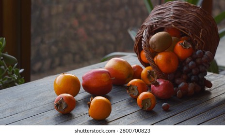 A high-angle diagonal composition of a rustic wicker basket on its side, with an abundance of fresh autumn fruits including red grapes, kiwi, oranges, ripe persimmons, apples, and a pomegranate spilling across a dark wooden patio table.                               