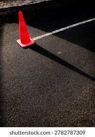 High-angle, dramatic shot of a bright orange traffic cone on dark asphalt, casting a long, sharp diagonal shadow next to a white parking line under harsh sunlight.