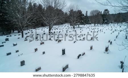 A high-angle drone photograph capturing an expansive cemetery covered in deep winter snow, with rows of granite headstones surrounded by dense pine trees on a cold, overcast day.