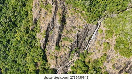 A high-angle drone shot captures a thin waterfall flowing down a steep, rocky cliffside.