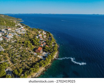 High-angle drone shot capturing charming coastal homes and villas built on a hillside. Houses are surrounded by lush green trees, overlooking a small bay and the clear Dalmatian Coast waters