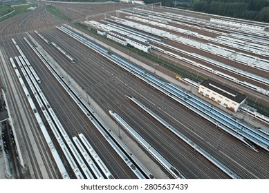 High-angle drone shot capturing a massive train yard. Streamlined high-speed trains sit on intricate rust-colored rails. Maintenance workshops and green vegetation frame this industrial hub, emphasizing advanced transport infrastructure and capacity.