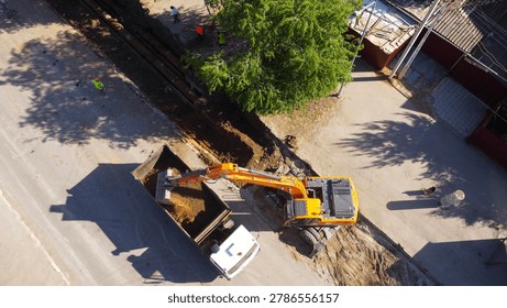 A high-angle drone shot capturing an orange hydraulic crawler excavator digging a trench and loading soil into the back of a white tipper dump truck