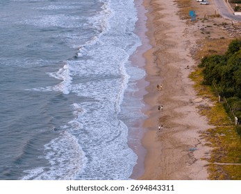 High-angle drone shot looking down on a sandy beach. People walk along the shore as white ocean waves break on the sand, next to dunes and beach amenities