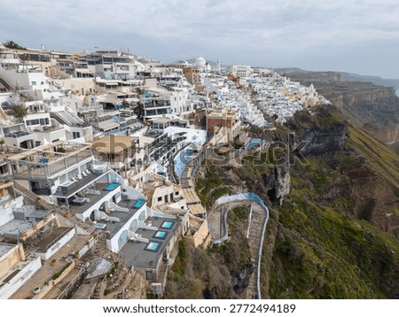 A high-angle drone shot looking down at the densely packed whitewashed hotels, infinity pools, and winding cliffside paths of Fira, Santorini, overlooking the volcanic caldera.