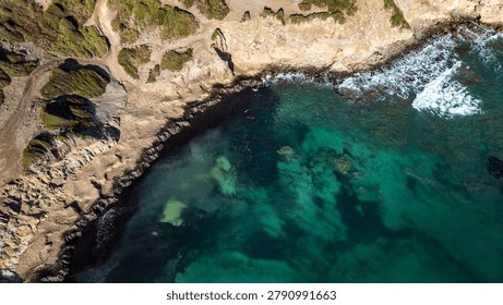 High-angle drone shot of a wild rocky coastline where transparent Mediterranean waters reveal a diverse seabed. The contrast between the warm earth tones, coastal scrub, and various shades of blue and emerald green creates a compelling and abstract natural composition.