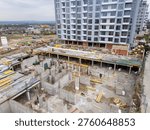 High-angle drone view capturing a complex construction site in progress. Focus is on the detailed groundwork of the lower level, with visible formwork, rebar, workers, and materials. A completed high-rise apartment building stands prominently in the background.