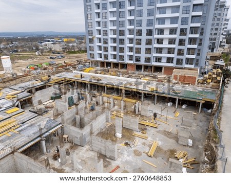 High-angle drone view capturing a complex construction site in progress. Focus is on the detailed groundwork of the lower level, with visible formwork, rebar, workers, and materials. A completed high-rise apartment building stands prominently in the background.