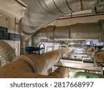 High-angle elevated view of a heavy industrial insulated piping network featuring a large blue gear-operated handwheel valve, metal-clad thermal insulation jacketing, and elevated steel catwalks inside a central mechanical plant room, illustrating HVAC chilled water distribution, thermal energy efficiency, and facility engineering infrastructure.