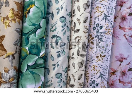 A high-angle full frame shot showcases six rolls of textile fabrics standing vertically side by side, creating a vibrant pattern composition. Each fabric features a distinct botanical and floral motif on a light background