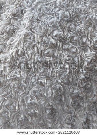 High-angle, full-frame close-up shot capturing the exquisite and intricate texture of a premium light grey faux fur fabric. The material features an elegant, stylized pattern of dense, swirling fibers that mimic the organic, delicate look of blooming rosebuds or whimsical coastal waves. Each individual synthetic thread catches the light softly, creating a beautiful interplay of subtle silver highlights, neutral grey midtones, and deep, velvety shadows within the crevices of the curls. The texture feels incredibly tactile, plush, cozy, and warm, evoking a sense of modern comfort, high-end interior design, and winter luxury. This seamless-looking textile background is perfectly suited for a wide variety of creative projects, including fashion backdrops, lifestyle branding, cozy home decor concepts, greeting cards, product staging, and abstract digital wallpapers. The neutral color palette ensures high versatility for graphic design overlay text or minimalist themes
