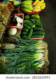 A high-angle, full-frame shot capturing a colorful and diverse assortment of fresh vegetables neatly arranged at a traditional open-air market stall. The foreground features large, bundled bunches of vibrant green scallions (green onions) with their fibrous white roots prominently displayed. Behind them, a pink plastic basket is filled to the brim with smooth, deep green cucumbers, while an adjacent basket holds cut white cabbage. In the upper section of the frame, a bright red tray displays fresh sweet corn with green husks alongside peeled yellow corn cobs, accompanied by fresh orange carrots and corn kernels in a blue bowl nearby. The composition beautifully captures the authentic atmosphere of a local Asian market grocery, making it a perfect creative asset for culinary blogs, healthy diet advertisements, supermarket promotional materials, and farming harvest features.