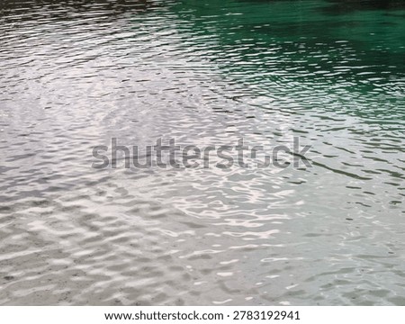 ​A high-angle, full-frame shot capturing the mesmerizing, gentle ripples on the surface of calm, clear water. The composition showcases a beautiful natural gradient, transitioning from a pale, sandy-toned transparency in the foreground to a deep, vibrant emerald green in the upper background. Sunlight reflects softly off the undulating wavelets, creating shimmering highlights and a textured, fluid pattern across the entire scene.