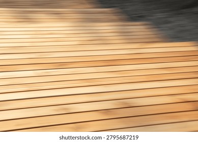 A high-angle, full-frame shot capturing a modern wooden boardwalk or decking area, utilizing an intentional camera movement (ICM) or panning technique to create a dynamic sense of motion and abstraction. The image features a series of parallel light-brown or tan wooden slats running horizontally across the frame. The intentional motion blur softens the natural wood grain texture into smooth, sweeping lines, stretching the warm golden tones of the wood from left to right. In the upper right corner, a dark, shadowy area contrasts sharply with the sunlit timber, adding a sense of depth and a geometric transition. The overall composition balances the rigid, linear pattern of the architecture with a fluid, artistic aesthetic, making it ideal as an abstract background, texture overlay, or conceptual design element representing speed, transition, or contemporary exterior design.