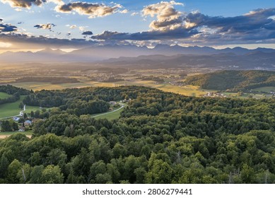 A high-angle landscape panoramic view over vast green forests, rolling hills, and sunlit farmlands in the Moravče valley at sunset, looking out from the vantage point of Sveta Trojica, Slovenia, toward the distant alpine peaks under evening sunbeams