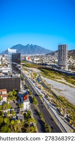 A high-angle landscape shot captures a dynamic corporate corridor in Monterrey, Mexico, under a solid, cloudless blue sky. In the foreground, contemporary glass skyscrapers and modern office complexes stand prominently along the main avenues. Cutting diagonally through the center of the scene is the expansive, dry gravel bed of the Santa Catarina River, bordered by green vegetation and active infrastructure construction sites. Multi-lane streets and urban transit routes flank the river corridor, leading the eye into the distant city sprawl. Dominating the background horizon is the majestic, sharp silhouette of the Cerro de la Silla mountain, framing this metropolitan center and illustrating the continuous vertical growth, structural evolution, and architectural modernization of this major Latin American financial hub.
