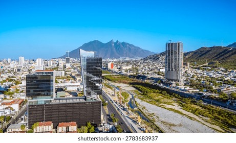 A high-angle landscape shot captures a dynamic corporate corridor in Monterrey, Mexico, under a solid, cloudless blue sky. In the foreground, contemporary glass skyscrapers and modern office complexes stand prominently along the main avenues. Cutting diagonally through the center of the scene is the expansive, dry gravel bed of the Santa Catarina River, bordered by green vegetation and active infrastructure construction sites. Multi-lane streets and urban transit routes flank the river corridor, leading the eye into the distant city sprawl. Dominating the background horizon is the majestic, sharp silhouette of the Cerro de la Silla mountain, framing this metropolitan center and illustrating the continuous vertical growth, structural evolution, and architectural modernization of this major Latin American financial hub.