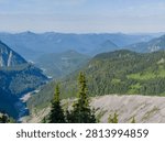 A high-angle landscape view of the Cascade Mountains in Washington state, featuring layers of blue ridges, a winding river valley, and dense evergreen forests.