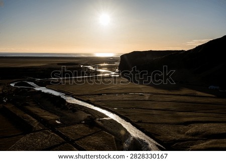 High-angle landscape view looking toward the Atlantic Ocean during golden hour, capturing a winding river reflecting the bright low sun across fields in southern Iceland.