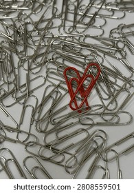 A high-angle macro shot featuring a heart-shaped arrangement of vibrant red paper clips centered amidst a scattered pile of metallic silver paper clips on a plain white surface. This image symbolizes love, focus, uniqueness, and finding the one among many.