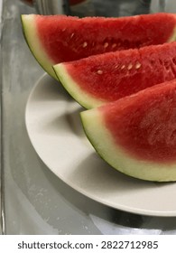 High-angle macro view of three vibrant, freshly sliced red watermelon wedges neatly arranged on a clean white plate, capturing the essence of healthy summer refreshment and natural vitality. The bright crimson flesh of the melon gleams with a natural juicy texture, highlighted by tiny light-colored seeds scattered throughout the segments, showcasing an appetizing and refreshing appearance. The thick, vibrant green rinds with their subtle pale borders provide a striking color contrast against the deep red interior and the neutral white plate background, creating a visually balanced and appealing composition.