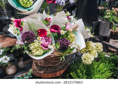A high-angle, medium close-up shot capturing a gorgeous selection of freshly assembled flower bouquets arranged inside a rustic woven wicker basket at a florist shop. The vibrant bouquets are elegantly wrapped in light-colored textured paper and feature a beautiful mix of blooms, including rich magenta calla lilies, deep purple dahlias, pink roses, light green carnations, delicate white daisies, and fluffy bunny tail grass. The display sits in a lush garden or flower shop setting, surrounded by other botanical arrangements, green foliage, and glass vases under soft, natural indoor lighting.