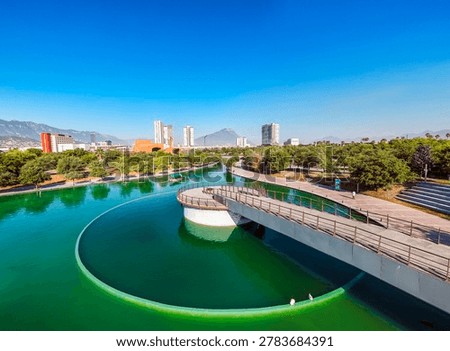 A high-angle medium landscape shot captures a modern concrete pedestrian bridge and circular overlook built directly above a wide section of the Paseo Santa Lucía canal in Monterrey, Mexico. The glistening turquoise water features a soft weir cascade on the left, while a small tour boat navigates the channel in the mid-ground. Elegant stone promenades and brick walking paths curve alongside the canal banks, shaded by rows of dense green trees. In the far background, the red brick structure of the Mexican Baseball Hall of Fame sits next to contemporary high-rise apartment towers and corporate buildings, framed by a distant mountain peak under a brilliant, cloudless blue sky, showcasing a premium urban recreational landscape.