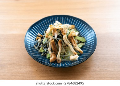 A high-angle, medium-range shot captures a vibrant Japanese salad served in a dark blue ribbed ceramic bowl, centered against a clean, warm-toned wooden tabletop. The dish is a refreshing and healthy medley of crisp bean sprouts, fresh cucumber, and dark seaweed, beautifully garnished with a mountain of golden-brown, crispy strips of deep-fried tofu (abura-age). The shallow depth of field keeps the fresh ingredients in sharp focus while softly blurring the background, emphasizing the appealing texture and bright colors of the meal. This inviting composition captures the essence of nutritious, modern home-style cooking, making it an excellent visual asset for food blogs, recipe websites, lifestyle magazines, or restaurant menu highlights.