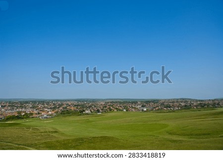 A high-angle panoramic view looking across the lush green fairways of Seaford Head Golf Course in East Sussex, UK. The vast landscape overlooks the coastal town of Seaford with its red-roofed houses, extending toward the horizon under an expansive, clear blue sky.