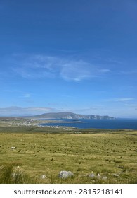 High-angle panoramic view overlooking green coastal pastures and a coastal village along a deep blue ocean bay with dramatic headlands under a vast sky