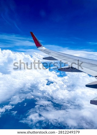 High-angle passenger window view of an airplane wing with a bright red winglet cruising high above a spectacular, thick layer of white cumulus clouds under a deep blue sky.