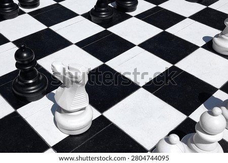 A high-angle perspective captures large, oversized chess pieces positioned on a classic outdoor board featuring alternating dark and light tiles. On the presented scene, a prominent white knight stands centrally, facing toward the left, while a formidable black bishop remains nearby, observing the tension across the monochromatic grid. The stark sunlight casts sharp, elongated shadows, adding depth and dimension to the expansive surface. Further pieces, including additional white pawns or figures, are scattered toward the bottom edge, creating a sense of an ongoing, strategic engagement in a public space. The texture of the square pavement tiles appears worn yet sturdy, grounding the monumental game elements in a real-world environment. Every sculpture exhibits smooth, rounded contours typical of traditional sets, yet their colossal scale transforms the ordinary surroundings into a theater of tactical decisions. The juxtaposition between the monochromatic flooring and the dark silhouettes creates a visually striking contrast, emphasizing the interplay of geometry and competition. As the light shifts, the dramatic shade lines shift alongside them, highlighting the tactile nature of these figures. The composition effectively focuses on the central horse-like statue, serving as the focal point of this intense, silent clash. Every detail, from the polished surfaces of the dark monoliths to the clean, bright finishes of the lighter counterparts, contributes to an atmosphere of focused deliberation and quiet intensity, capturing a singular, frozen moment of intellectual challenge where every move carries significant weight on the checkered landscape.