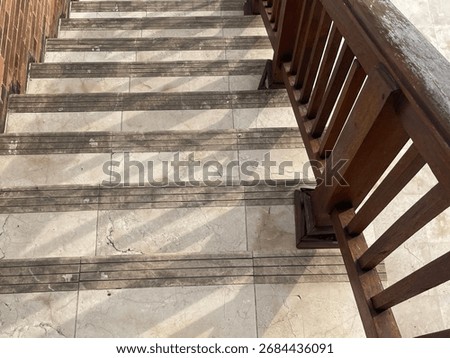 A high-angle perspective looking down a long, weathered stone staircase with a dark wooden railing. The scene evokes a sense of history, architecture, and a pathway leading to a tiled courtyard.