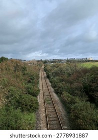 A high-angle, perspective shot looking down a single-track railway line in Pembrokeshire, Wales. The iron rails, bedded in grey gravel ballast with patches of green moss, stretch into the distance through a corridor of dense, wild gorse and autumn-toned bushes. In the background, the skyline of the historic seaside town of Tenby is visible, featuring the prominent tall spire of St Mary's Church and a row of colorful buildings along the cliffside. The scene is captured under a heavy, dramatic overcast sky, typical of the Welsh coast, looking toward the town from a bridge along the Pembroke Dock branch line.