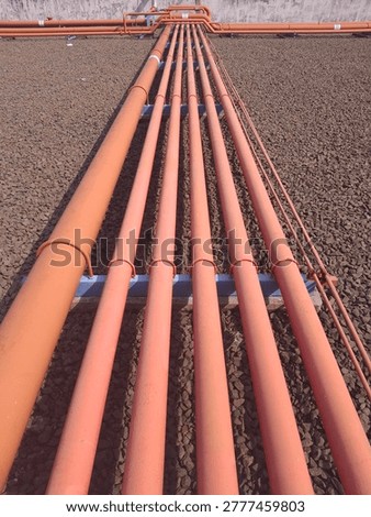 ​A high-angle perspective shot of several orange industrial pipes arranged in parallel on a gravel surface. The pipes are supported by blue metal frames, showing a clean and organized outdoor industrial infrastructure