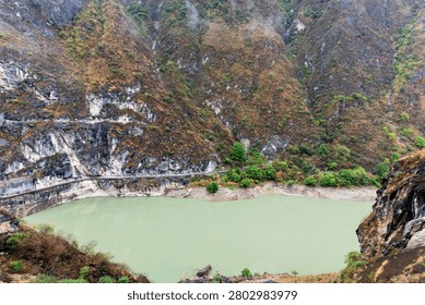 High-angle scenic view looking down into the dramatic bend of the Jinsha River canyon in Yunnan, China. A long, engineered cliffside walkway follows the natural curve of the sheer, heavily textured rock wall right above the calm turquoise water. The sheer drop-offs and towering canyon heights emphasize the thrilling, rugged nature of this legendary inland waterway.