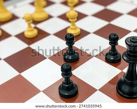 A high-angle, selective focus shot of black chess pieces positioned on a classic red and white checkered board. The image highlights the queen and pawns in the foreground while the opposing white pieces remain softly blurred in the background.