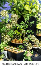 A high-angle selective focus shot of an elegant banquet table served with various gourmet appetizers, tartlets, and canapes adorned with lush green floral decorations outdoors