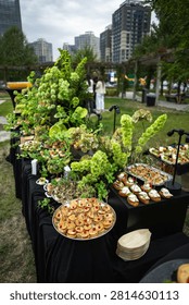 A high-angle selective focus shot of an elegant banquet table served with various gourmet appetizers, tartlets, and canapes adorned with lush green floral decorations outdoors