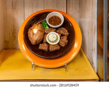 A high-angle shot capturing a detailed plastic food sample (shokuhin sampuru) replica of a Japanese diced beef fillet tenderloin steak (hire koro steak) platter displayed on a stand inside a restaurant showcase. The dish features tender cut steak pieces topped with a lemon wheel and herb butter, accompanied by a baked potato with butter, broccoli, and a small white ramekin filled with Japanese-style soy onion sauce (wafu onion sauce). Presented in a dark brown ceramic dish set inside an orange rimmed plate, this image is ideal for stock media on Japanese food models, family restaurant menus, yoshoku cuisine, and culinary displays.
