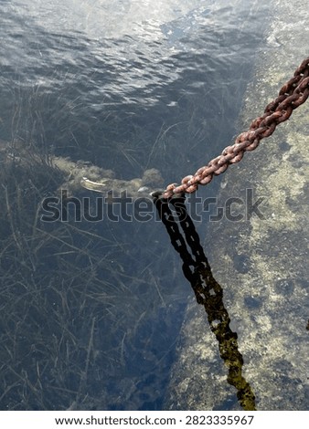 A high-angle shot capturing a heavy, rusty metal mooring chain extending from the rocky shore into the clear, transparent sea water. The texture of the sunlit ripples on the water surface contrasts with the dark, submerged links of the chain resting near the seabed. This image illustrates maritime infrastructure, coastal anchoring systems, and the rustic charm of a sea harbor or wild rocky beach.
