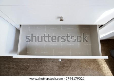   A high-angle shot looking directly down into an empty, clean drawer of a white wooden chest. The drawer features a light grey patterned liner and simple silver knobs, positioned over a tan carpeted floor.          