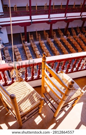 A high-angle shot looking down from the upper gallery, focusing on two foreground wicker chairs next to a red railing, with the lower patio seating visible below.