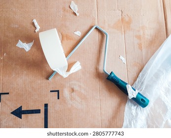 A highangle shot of a paint roller frame and masking tape on protective cardboard, capturing the messy, realistic process of DIY home improvement, renovation, and wall painting prep.