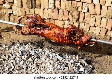 A High-angle Shot Of A Pig On A Spit Rotating Over The Hot Ashes On A Sunny Day Outdoors