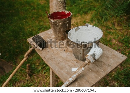 A high-angle shot of a rustic wooden workbench outdoors, holding weathered metal buckets filled with white and red paint. Old paintbrushes with textured bristles rest on the paint-splattered surface against a blurred green grass background.