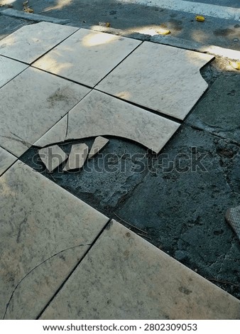 A high-angle shot showing shattered and missing pieces of light-colored outdoor floor tiles, exposing the rough dark concrete base underneath.