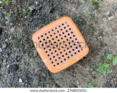 High-angle shot of a small, orange plastic stool with a perforated square seat placed on a rough, earthy ground outdoors.