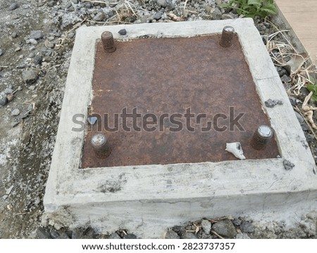 A high-angle shot of a square concrete foundation featuring a weathered rusty steel baseplate with four protruding anchor bolts.
