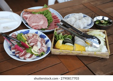 A high-angle shot of a table spread featuring plates of raw beef, pork, and chicken, accompanied by a bamboo basket filled with sliced vegetables like corn, cabbage, and eggplant. Tongs are placed on the plates and in the basket, suggesting an outdoor or indoor grilling experience. Bright, clean, and appetizing, this image is perfect for culinary blogs, meal preparation concepts, or barbecue event marketing.